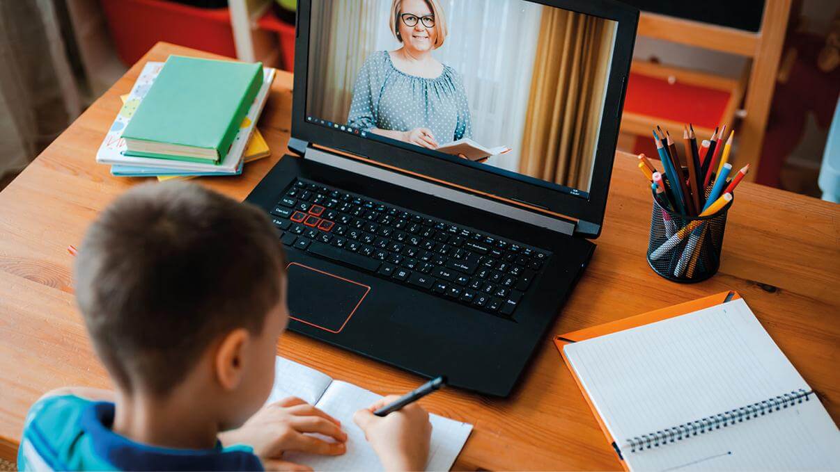 Un bambino è seduto a una scrivania con un quaderno e una penna in mano, guardando uno schermo di laptop dove appare una persona che sembra insegnare o spiegare qualcosa. Sulla scrivania ci sono anche alcuni libri impilati di lato e un quaderno aperto con contenuti scritti accanto al laptop. È visibile anche una tazza piena di matite colorate. Questa scena probabilmente rappresenta una situazione di apprendimento a distanza, che è diventata più rilevante a causa dell’aumento dell’istruzione online.