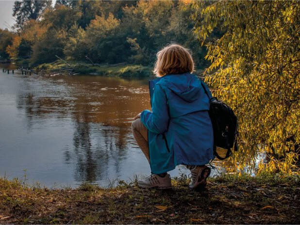 Persona con giacca blu e zaino nero accovacciata vicino a un fiume, osservando l'acqua e circondata da alberi in una giornata autunnale.