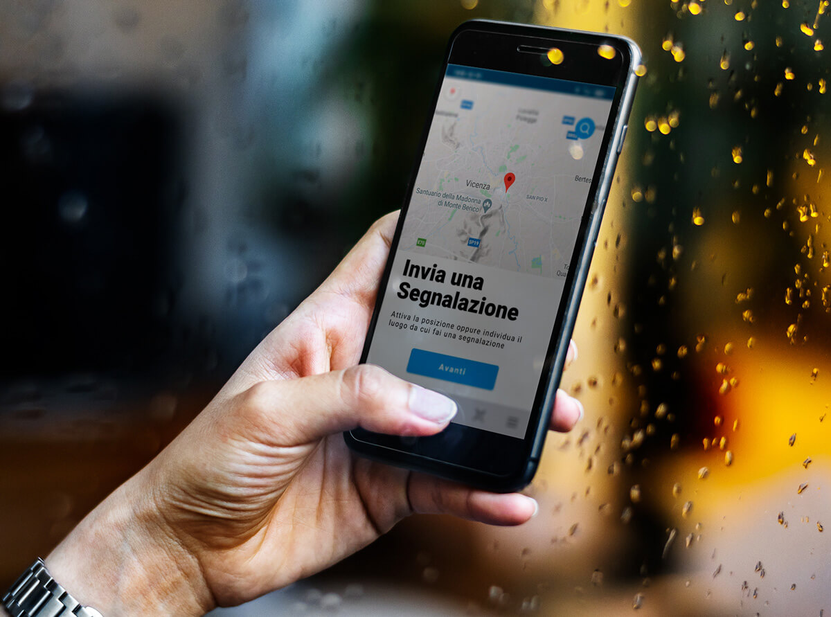 Una mano tiene uno smartphone che mostra un'applicazione in italiano per inviare segnalazioni geolocalizzate. Sullo sfondo, una finestra con gocce di pioggia.
