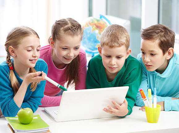 Quattro bambini seduti a un tavolo guardano insieme lo schermo di un laptop bianco, sorridendo e conversando.