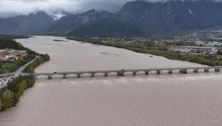 Vista aerea di un fiume in piena che scorre sotto un ponte ad arcate in una zona montuosa. Il fiume è di colore marrone a causa del volume dell'acqua e del sedimento trasportato. Il ponte collega due rive con edifici e vegetazione, mentre sullo sfondo si vedono montagne coperte da nuvole scure.