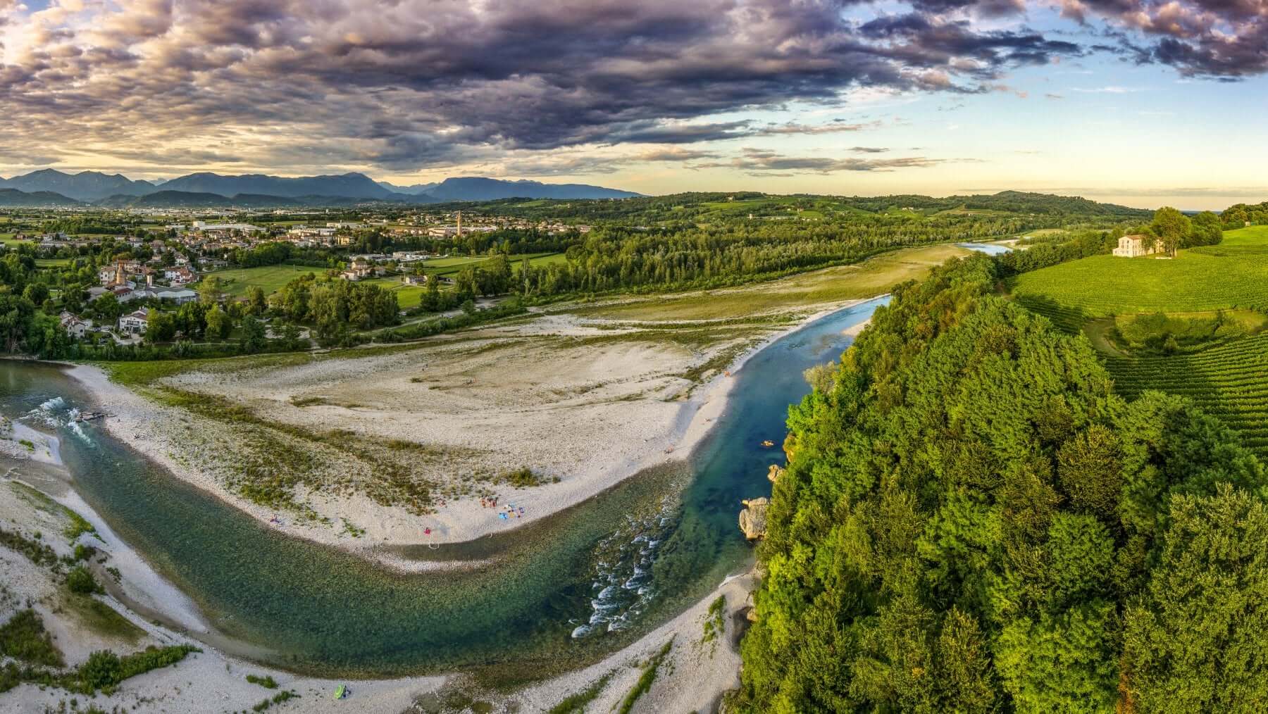 Panorama aereo di un fiume che serpeggia attraverso un paesaggio verde. In primo piano, una spiaggia di ghiaia forma un'ampia curva. Sullo sfondo, colline boscose, campi coltivati e un piccolo centro abitato. Cielo drammatico con nuvole al tramonto.